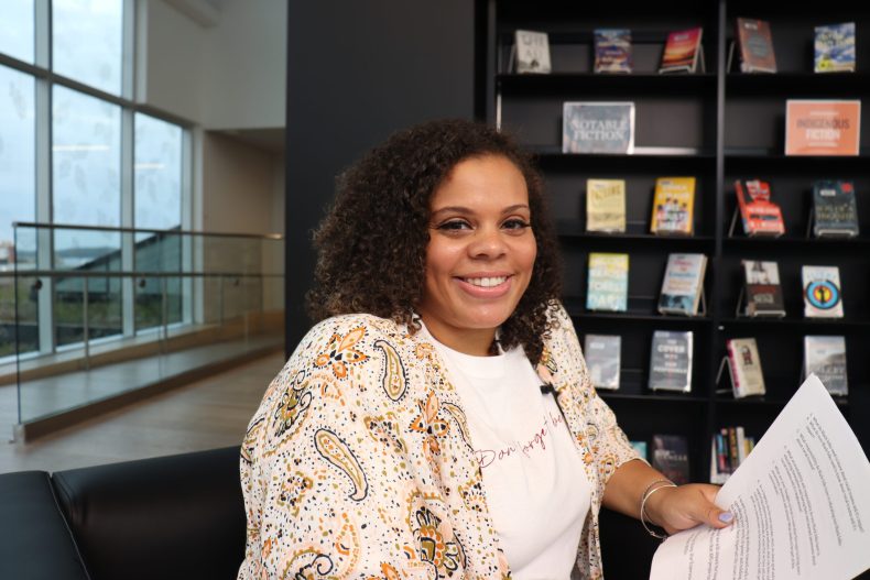 Black Lady with light coloured clothing smikes for the camera with books on a bookcase in the background.